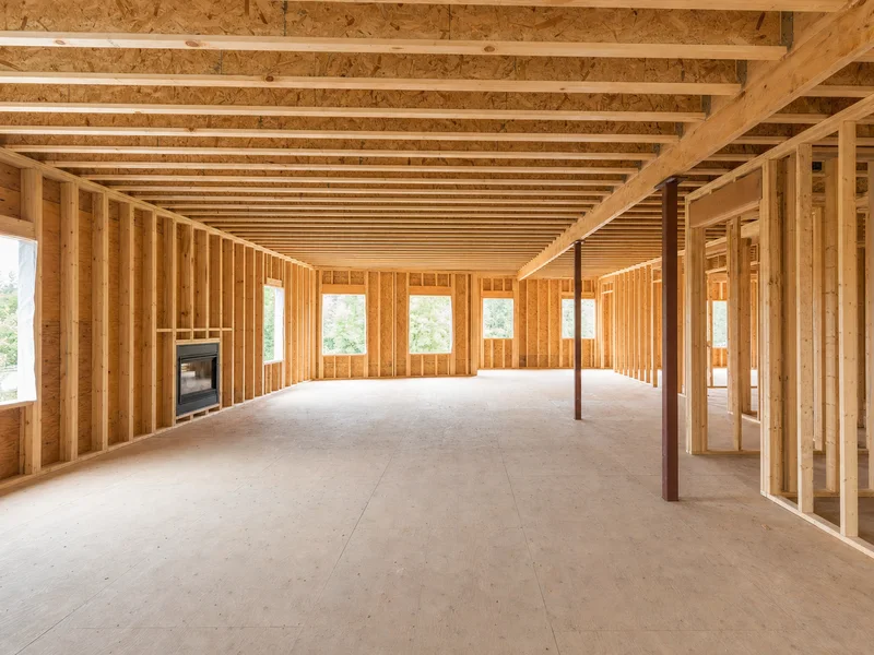 A house under construction: bare stud framing, plywood subfloor, and a zero-clearance fireplace already set into the wall on the left