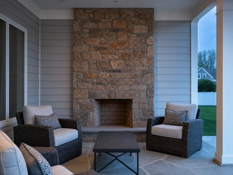A stone outdoor fireplace on a covered back patio at dusk with wicker seating